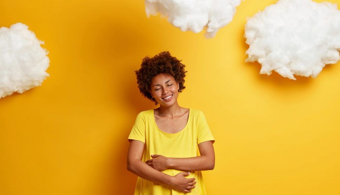 Pleased African American woman embraces pregnant belly, expresses love for unborn child, smiles happily, enjoys last months of pregnancy, isolated on yellow wall. Expectant mother hugs tummy