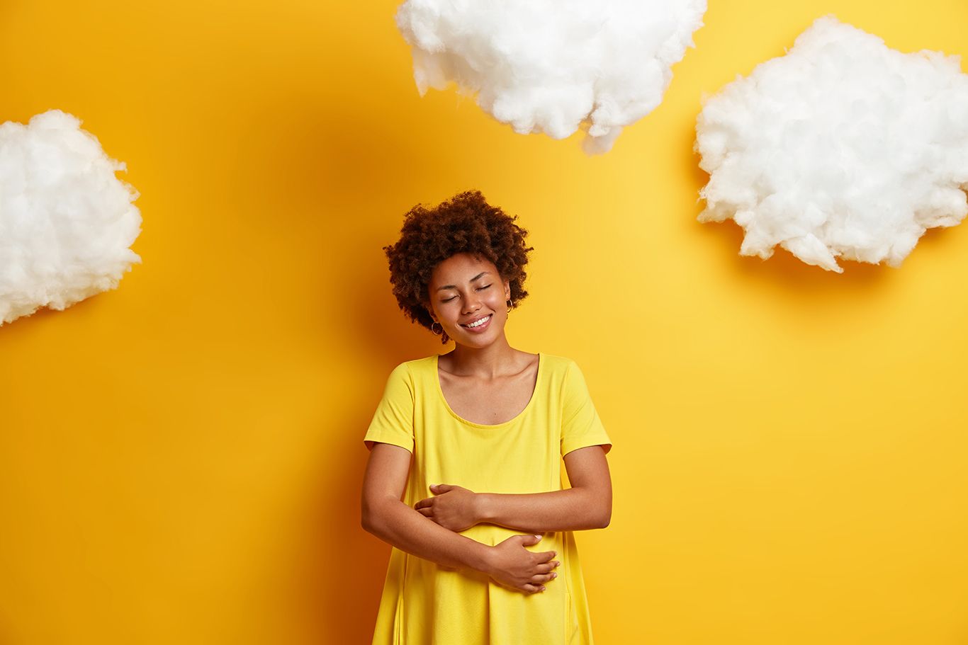 Pleased African American woman embraces pregnant belly, expresses love for unborn child, smiles happily, enjoys last months of pregnancy, isolated on yellow wall. Expectant mother hugs tummy
